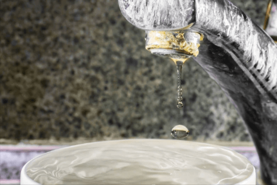 An old faucet with calcium buildup on it, dripping water into a full glass of water.