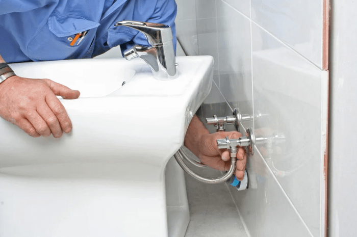 A plumber in a blue uniform is installing a new faucet and a white ceramic sink in a commercial restroom.