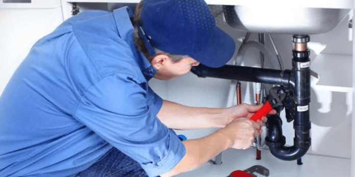 A plumber in a blue uniform and hat fixing a sink drain with a wrench.