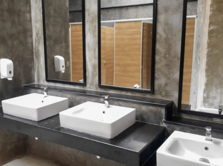 A row of wide square commercial faucets and white ceramic sinks set in black granite counters against a brown-grey stone cement wall.